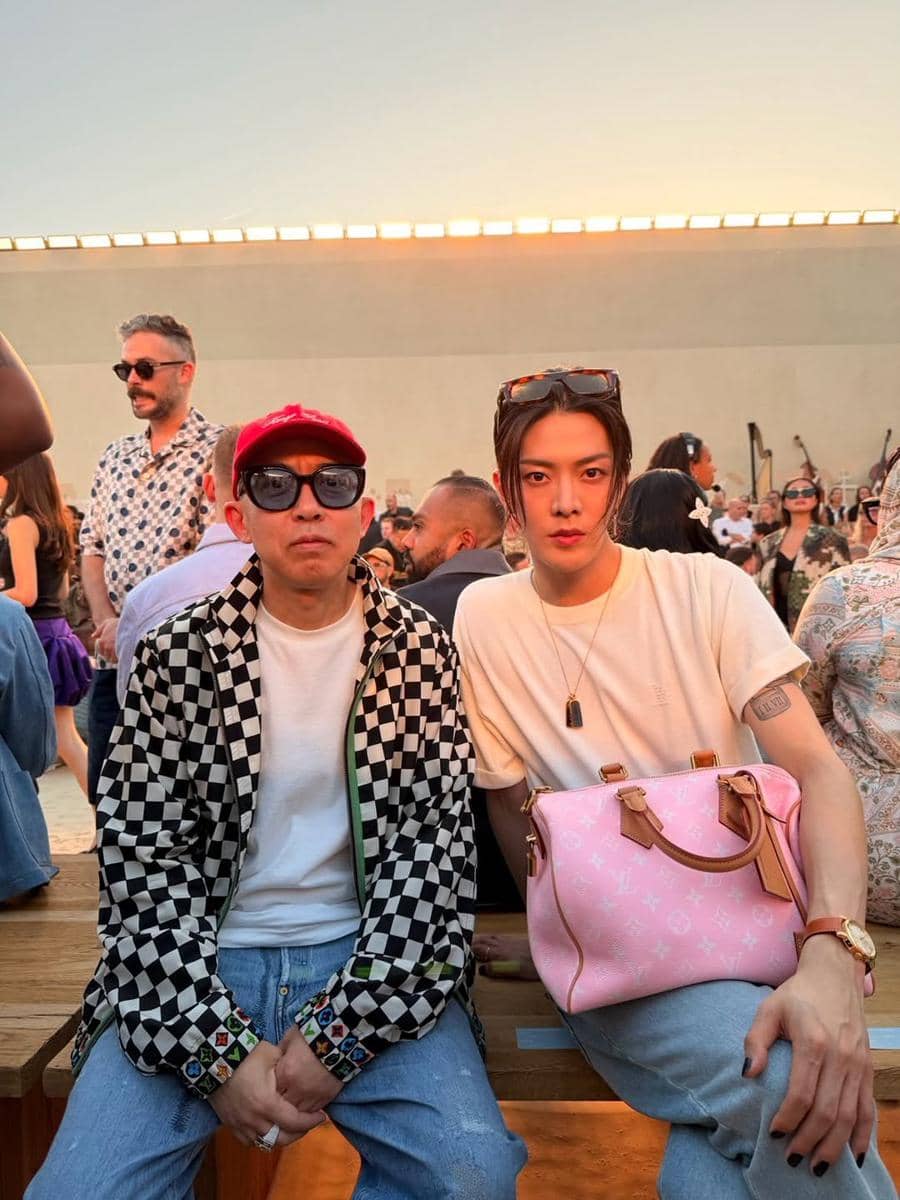 Dua pria duduk di acara luar ruangan saat matahari terbenam, salah satunya mengenakan kaus putih dan tas pink, sementara yang lain memakai jaket motif kotak-kotak.