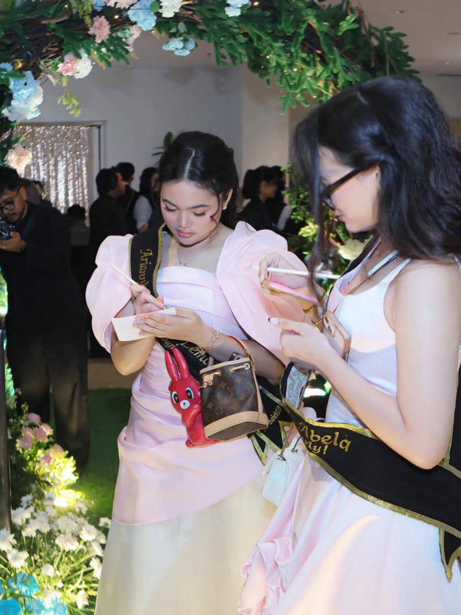 Dua remaja mengenakan gaun berwarna pink muda dengan selempang hitam emas sedang menulis di kartu pada acara prom night sekolah.