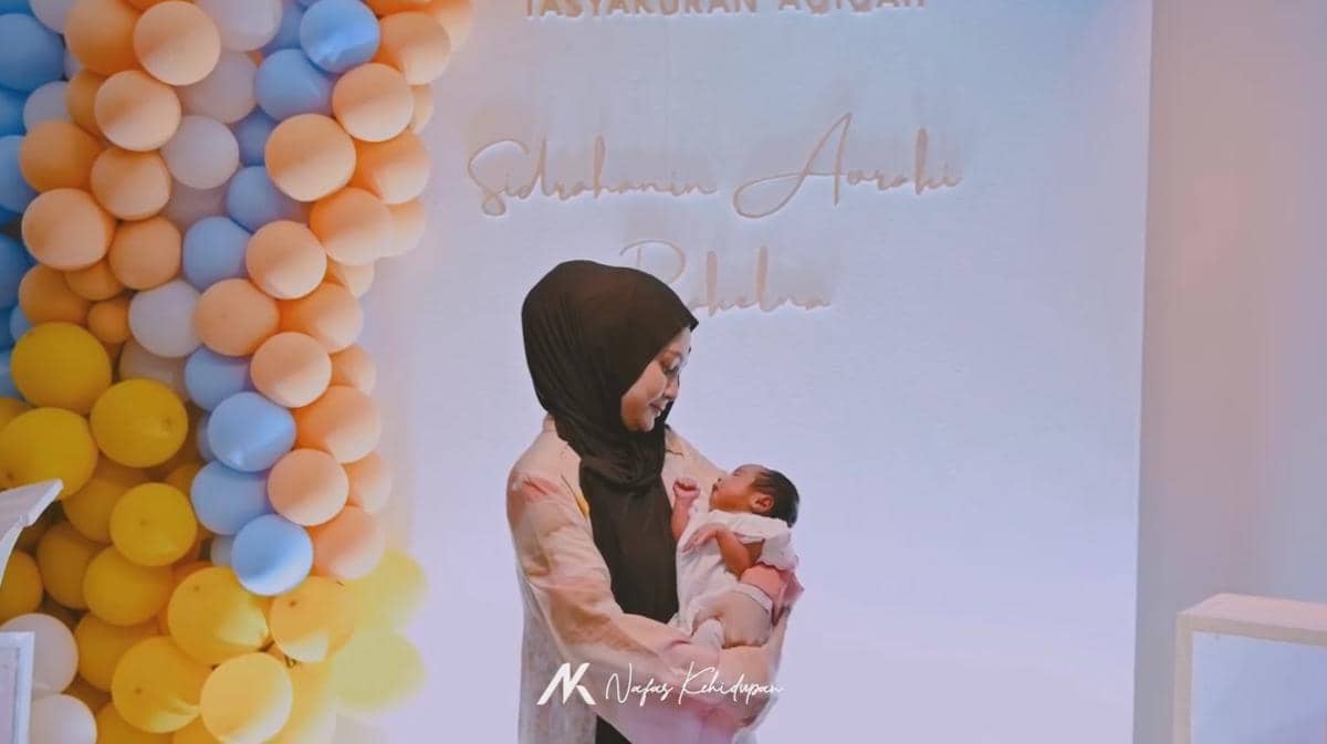 Seorang wanita berhijab memegang bayi di acara akikah dengan latar dekorasi balon warna kuning, biru, dan oranye pastel.