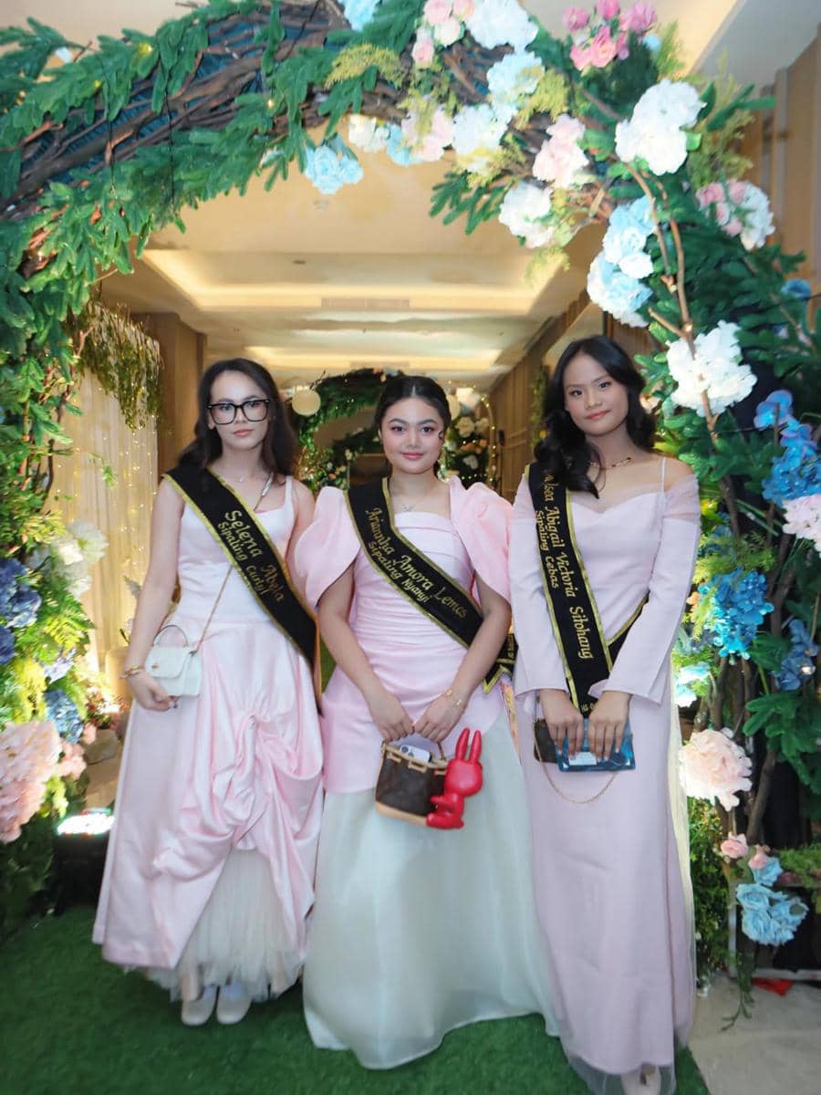 Tiga remaja mengenakan gaun berwarna pink muda dengan selempang hitam berpose di bawah dekorasi bunga pada acara prom night SMP.