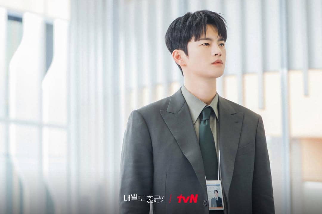 Seorang pria mengenakan setelan jas abu-abu dan dasi hijau berdiri di dalam gedung modern dalam cuplikan drama See You at Work Tomorrow.
