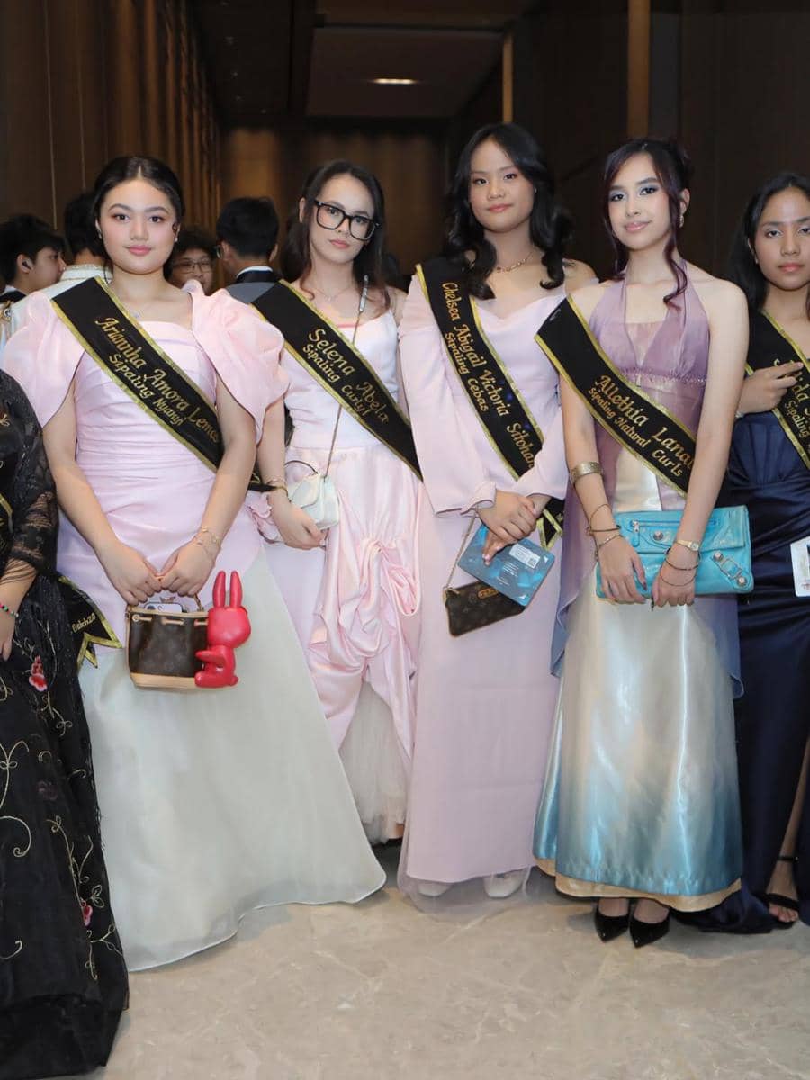 Sekelompok remaja perempuan mengenakan gaun elegan dan selempang hitam beraksen emas saat menghadiri acara prom night sekolah.