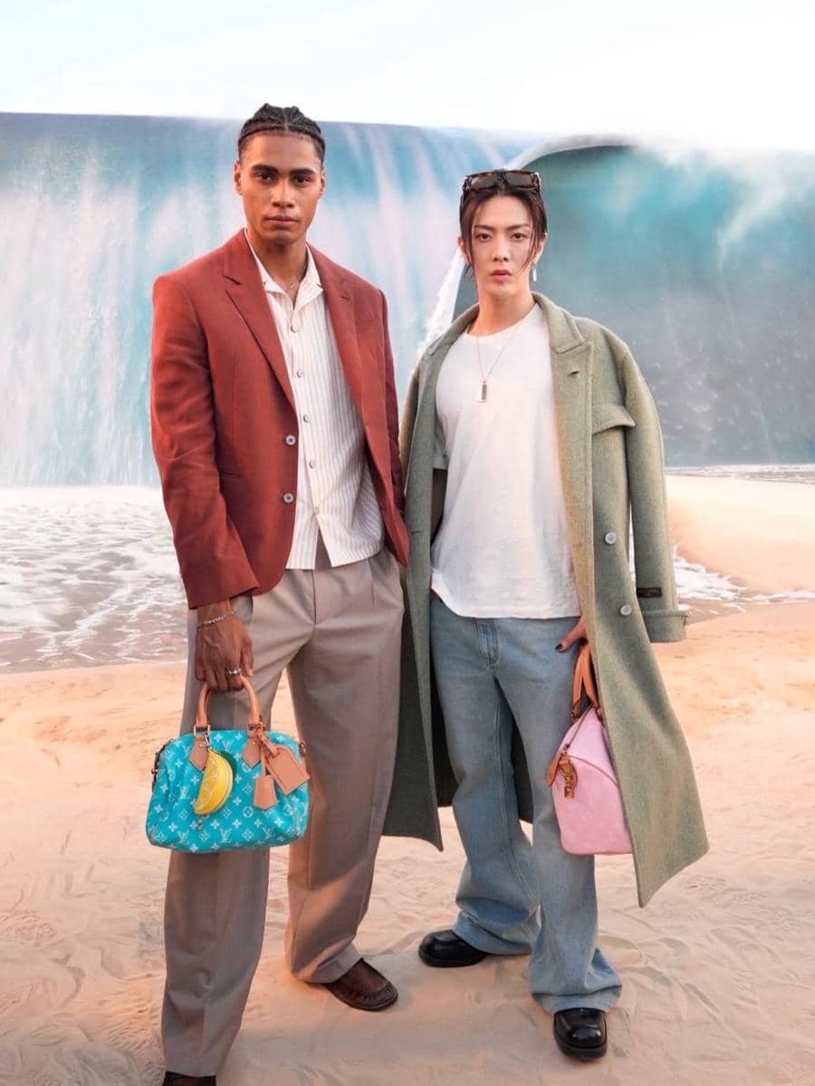 Dua pria berpose di tepi pantai buatan dengan latar ombak besar, mengenakan busana bergaya modern dan membawa tas tangan warna cerah.