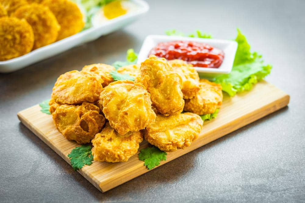 Beberapa potong bakwan tahu goreng berwarna keemasan disajikan di atas talenan kayu dengan saus sambal di sampingnya.