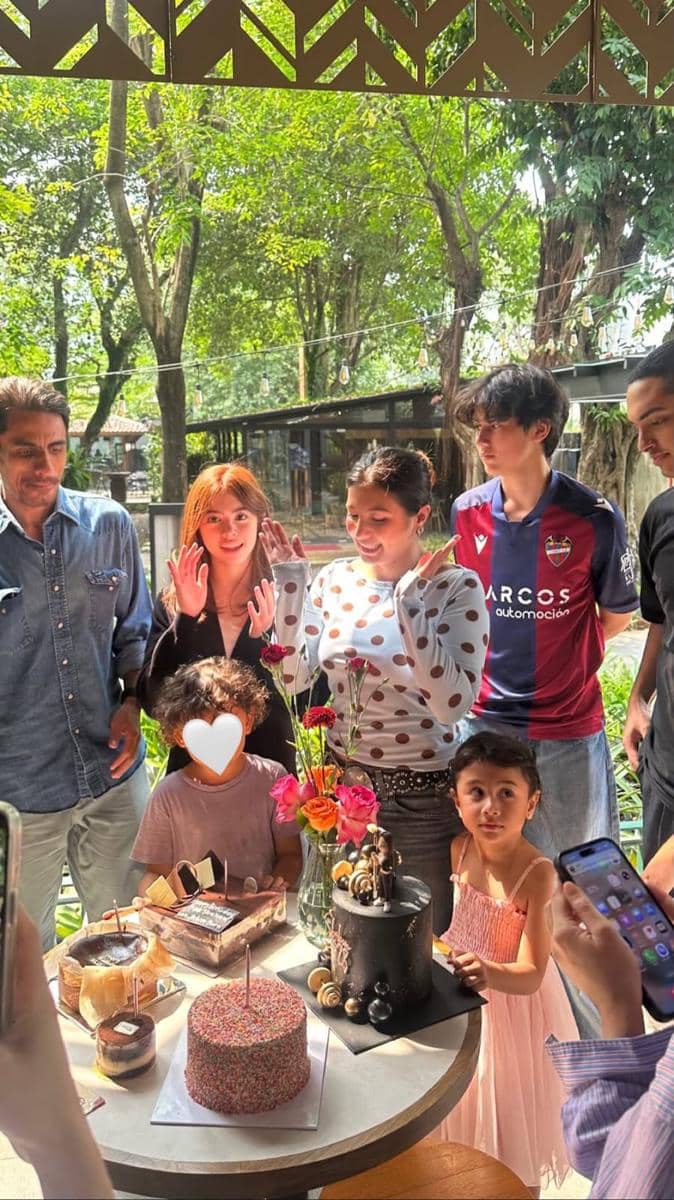 Sekelompok orang merayakan ulang tahun di luar ruangan dengan beberapa kue di atas meja dan suasana hangat di bawah pepohonan.