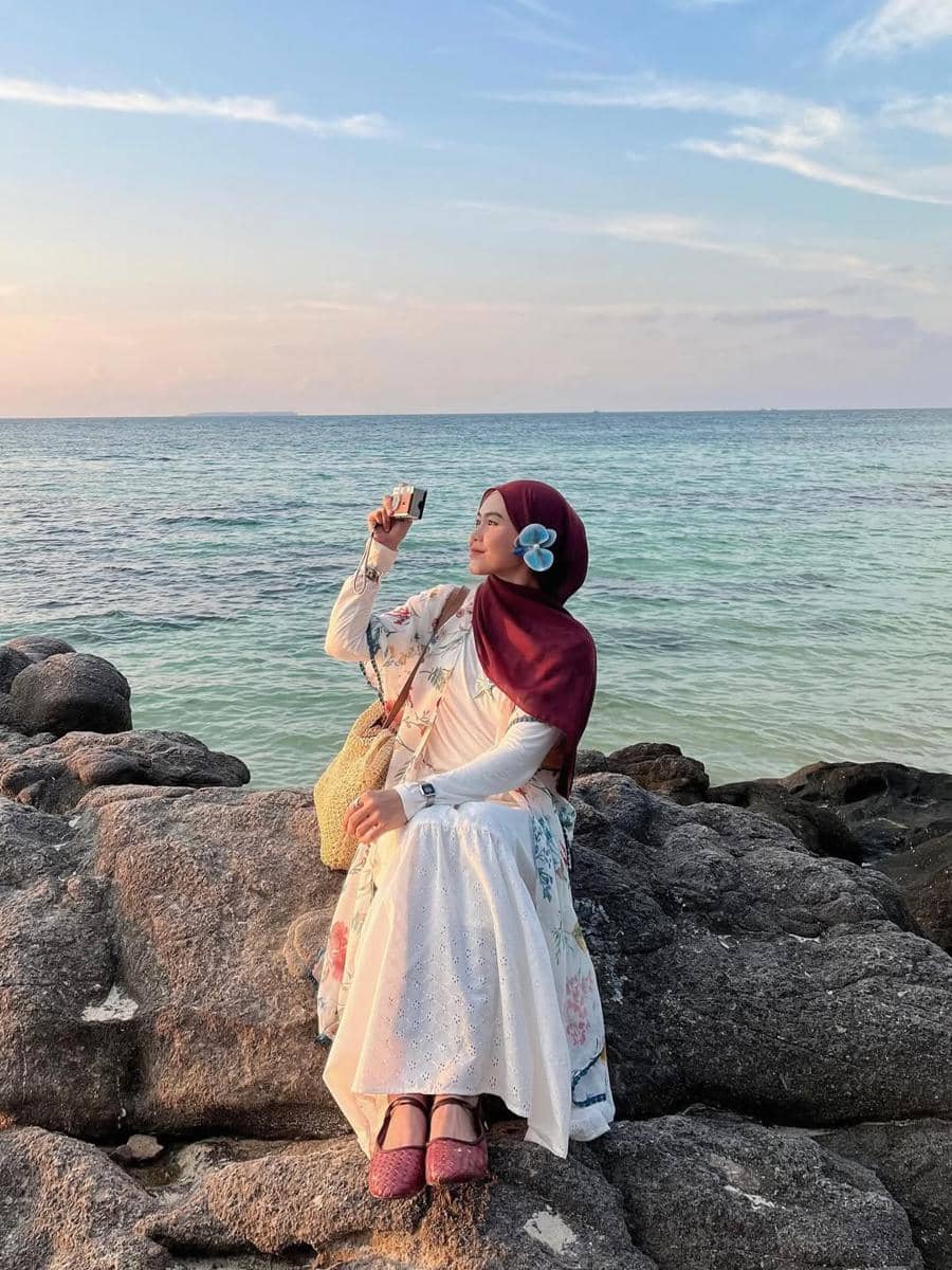 Seorang wanita berhijab duduk di atas batu tepi pantai saat matahari terbenam, mengenakan gaun putih bermotif bunga dan hijab marun.