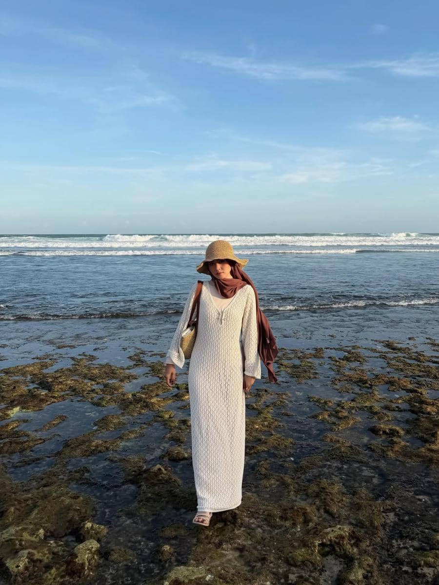 Seorang wanita mengenakan gaun panjang putih dan topi jerami berjalan di tepi pantai dengan latar ombak dan langit biru cerah.