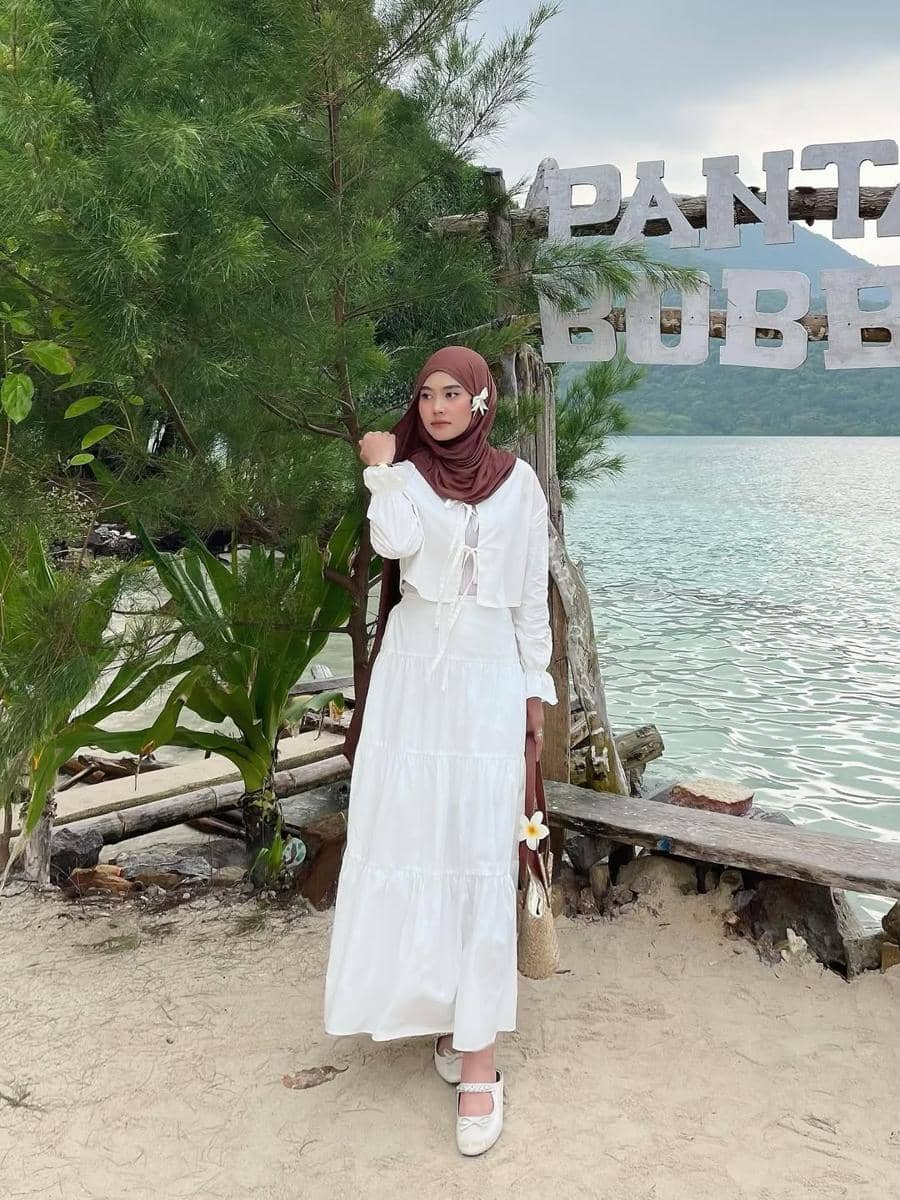 Seorang wanita berhijab cokelat mengenakan gaun putih panjang berdiri di tepi pantai dengan latar laut dan pepohonan hijau.
