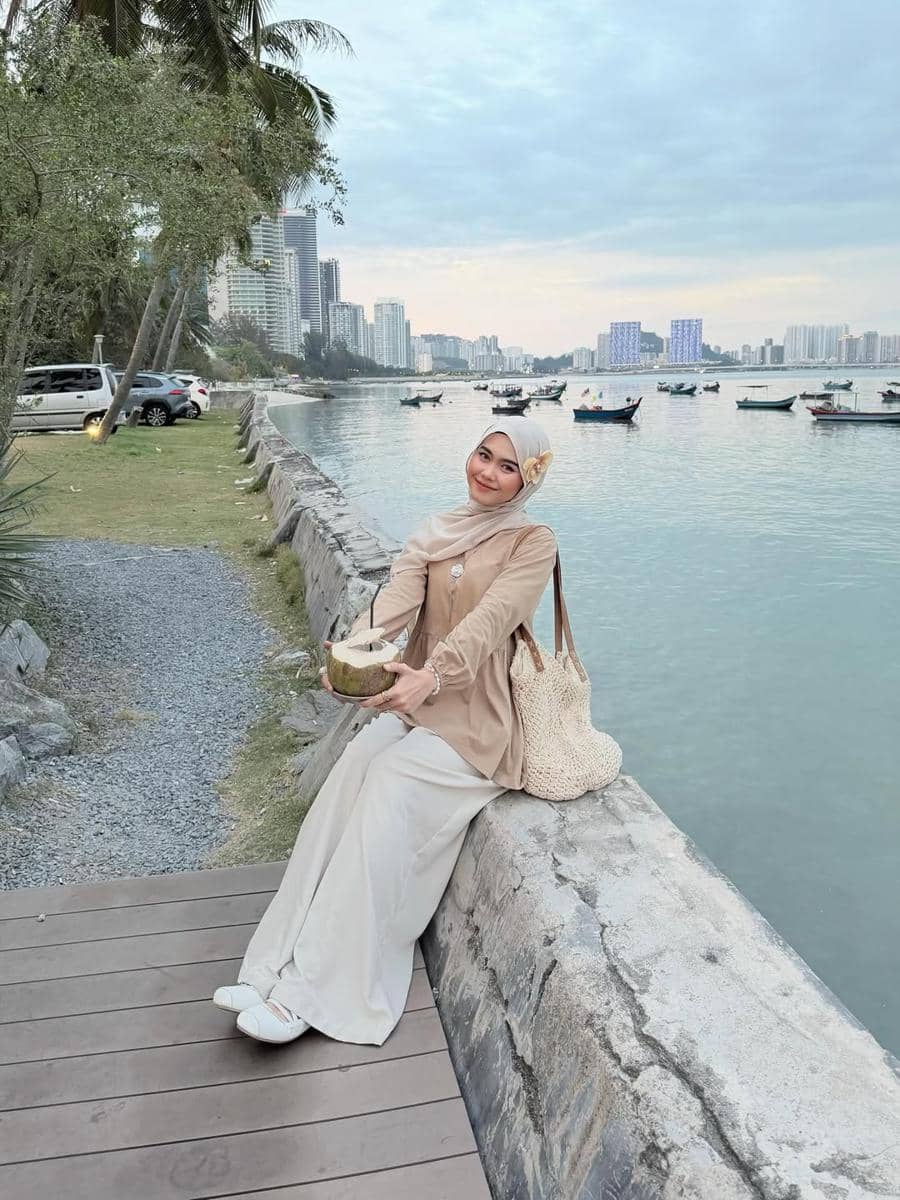 Seorang wanita berhijab duduk di tepi pantai sambil memegang kelapa muda, dengan latar gedung tinggi dan perahu di laut tenang.