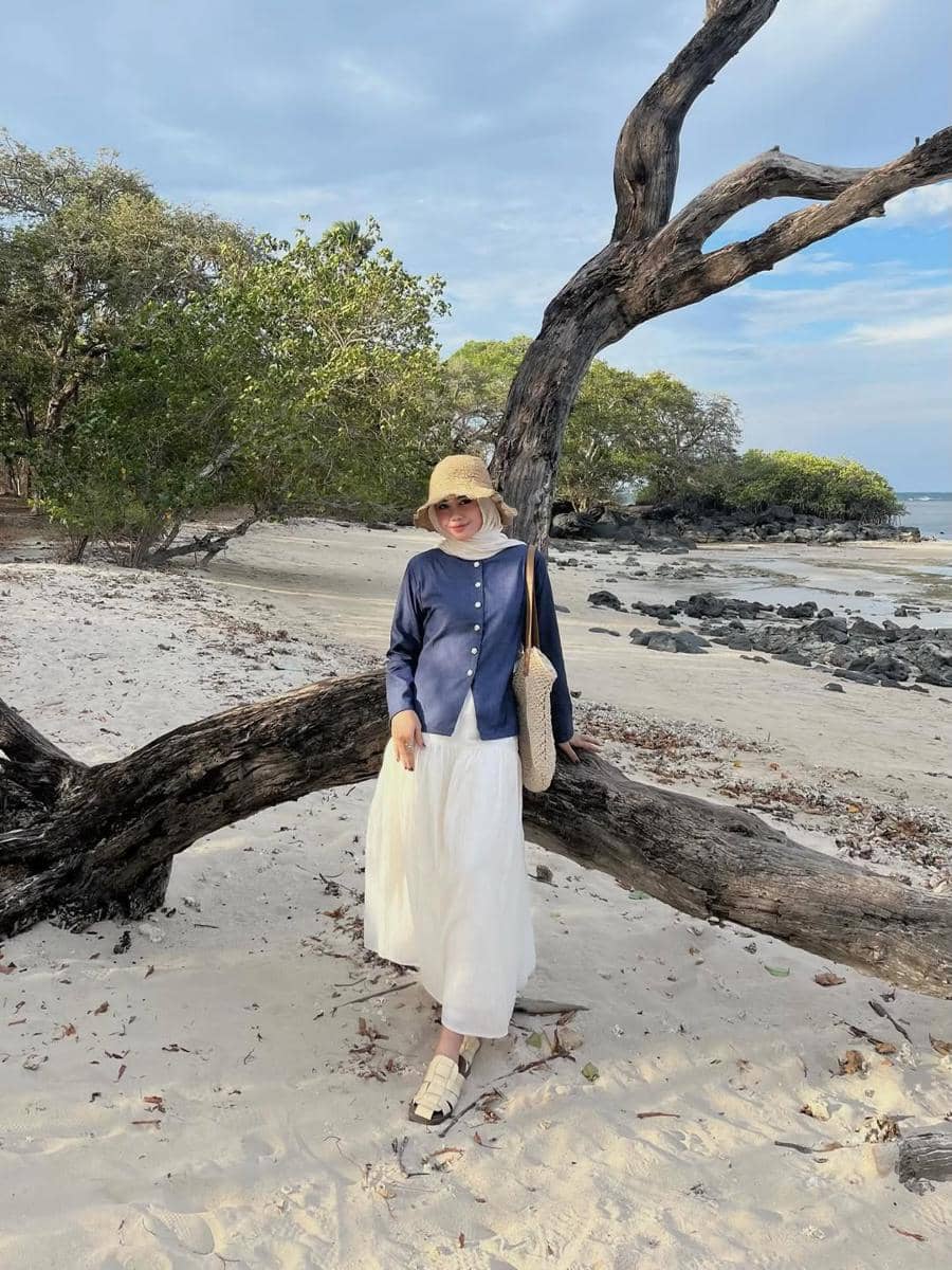 Seorang wanita berhijab mengenakan topi jerami, atasan biru, dan rok putih berpose di pantai berpasir dengan pohon kering dan latar laut.