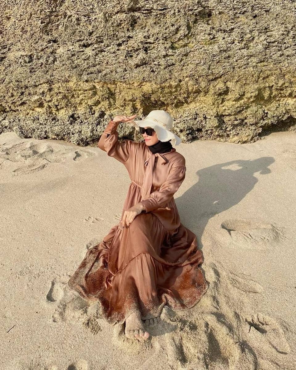 Seorang wanita berhijab mengenakan gaun panjang berwarna cokelat dan topi putih duduk di pasir pantai dengan latar tebing batu.