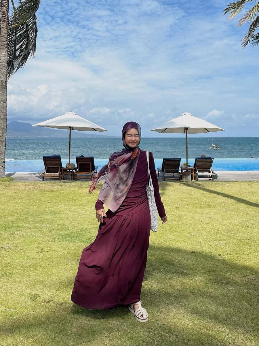 Seorang wanita berhijab mengenakan dress ungu berjalan di area rumput dekat pantai dengan latar kursi santai dan payung putih.