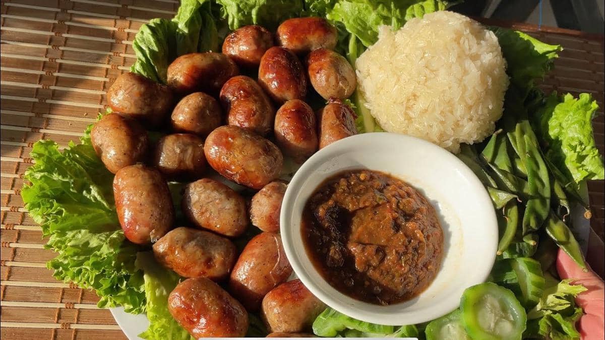 Sajian sai oua khas Laos disajikan dengan ketan, saus sambal pedas, dan sayuran segar di atas daun selada.