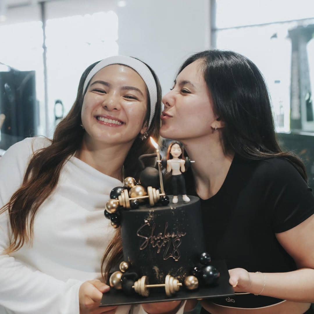 Dua wanita tersenyum sambil memegang kue ulang tahun hitam bertema olahraga dengan lilin dan hiasan figur kecil di atasnya.