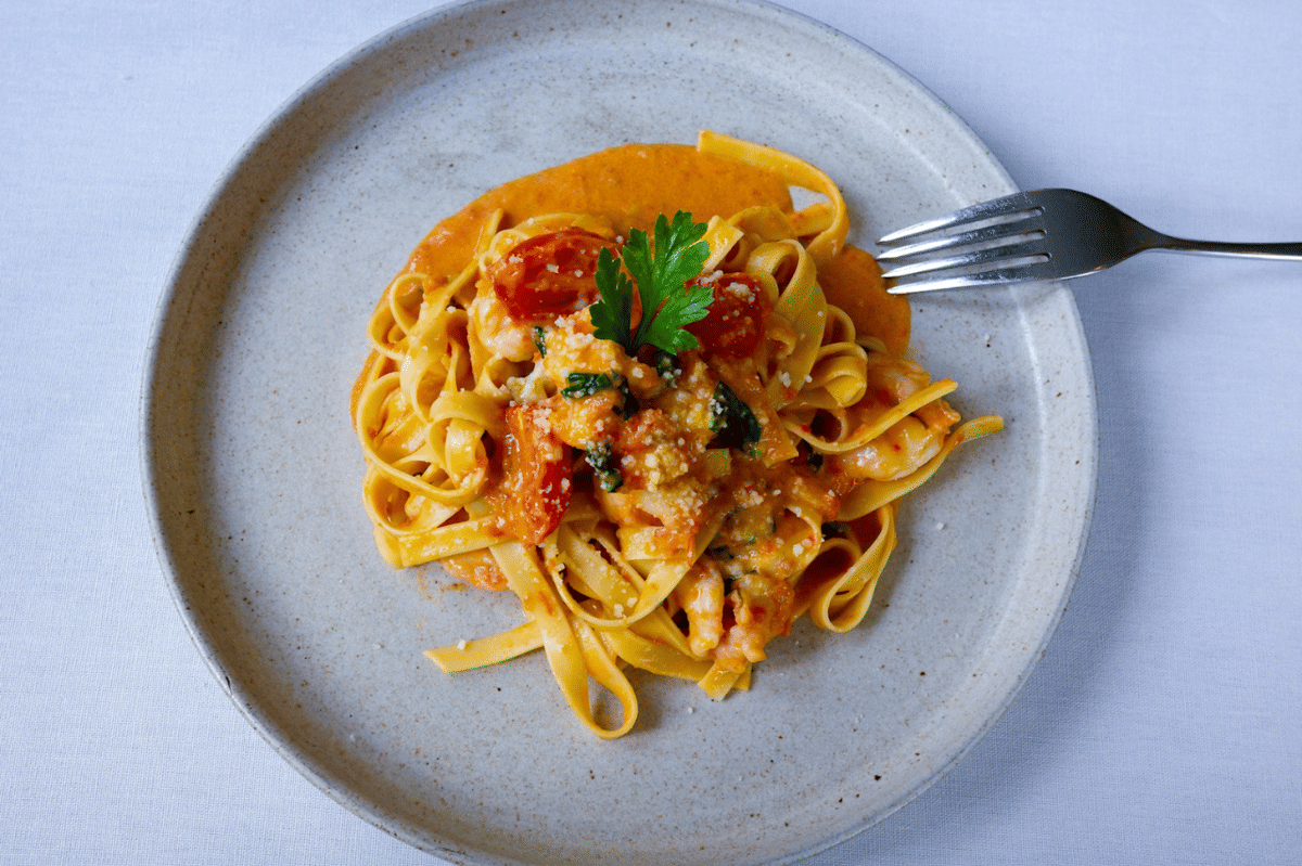 Sepiring tagliatelle dengan saus tomat dan 'nduja, dihiasi daun peterseli di atas piring keramik abu-abu muda dengan garpu di sampingnya.