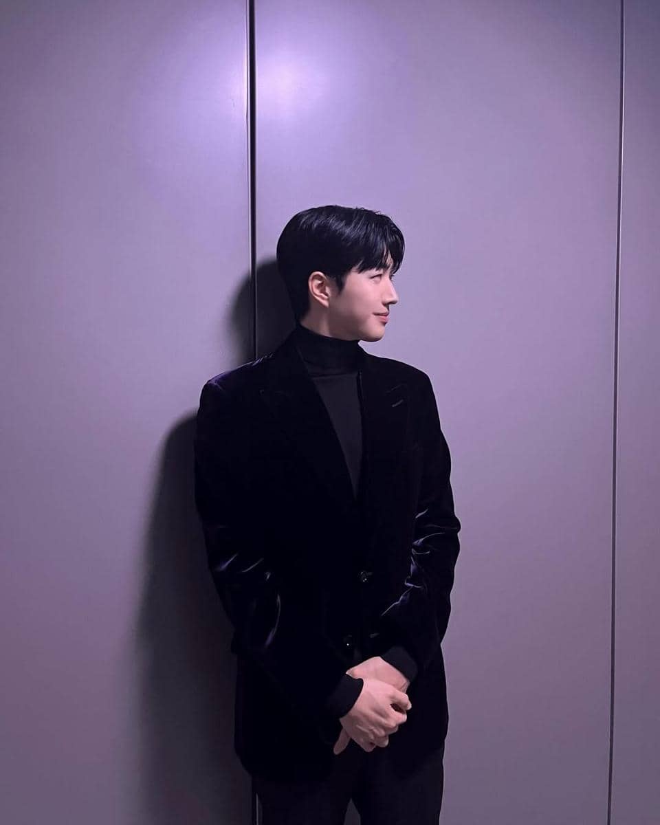 Seorang pria berambut hitam mengenakan setelan jas hitam dan kaus turtleneck berdiri di depan dinding berwarna ungu lembut.
