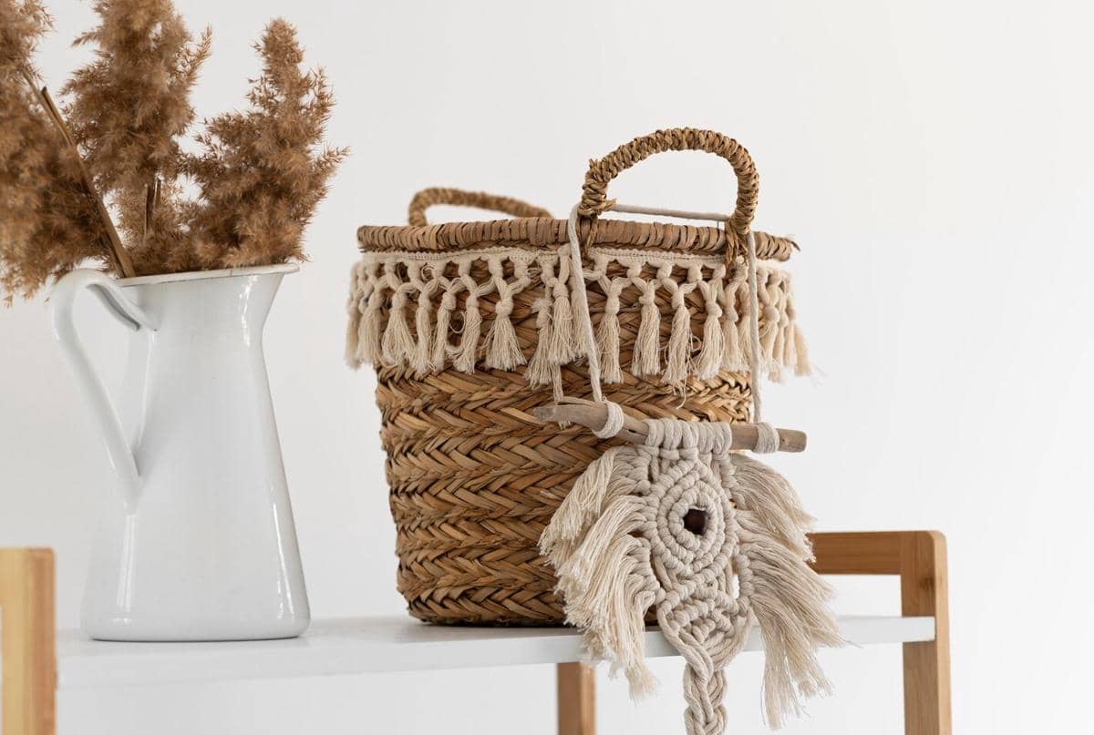 Keranjang anyaman rotan dengan hiasan tali makrame berwarna krem di atas meja putih, berdampingan dengan vas putih berisi tanaman kering.