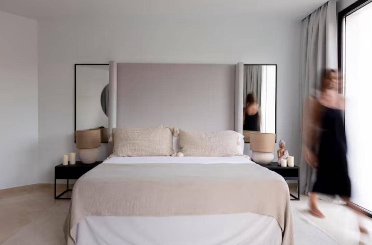 Kamar tidur bergaya minimalis dengan dominasi warna netral, tempat tidur besar, dua lampu meja, dan tirai abu-abu di dekat jendela besar.