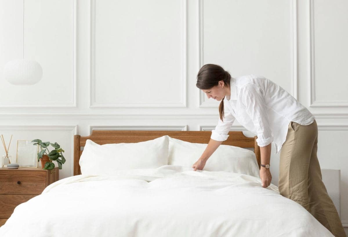 Seorang wanita mengenakan kemeja putih sedang merapikan tempat tidur dengan seprai putih di kamar bergaya minimalis.