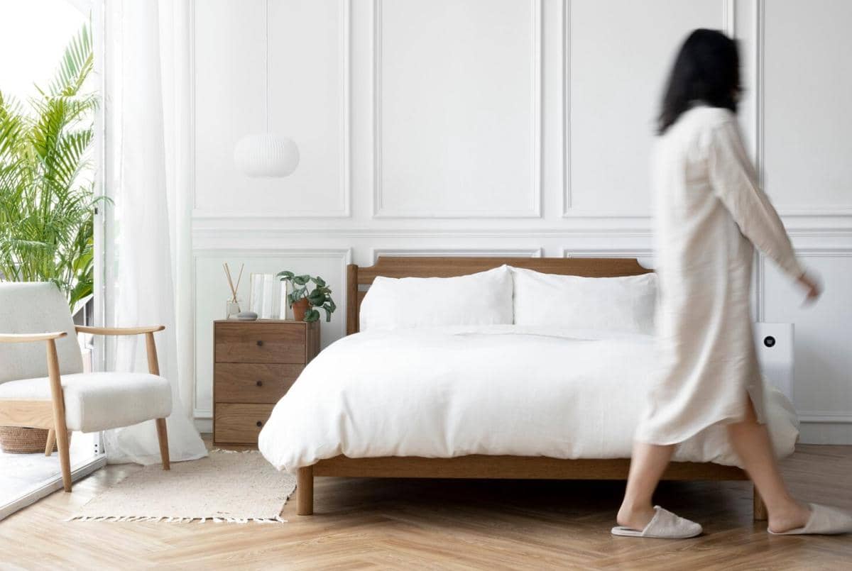 Kamar tidur minimalis dengan tempat tidur kayu, seprai putih, meja samping, dan tanaman hias di dekat jendela besar.