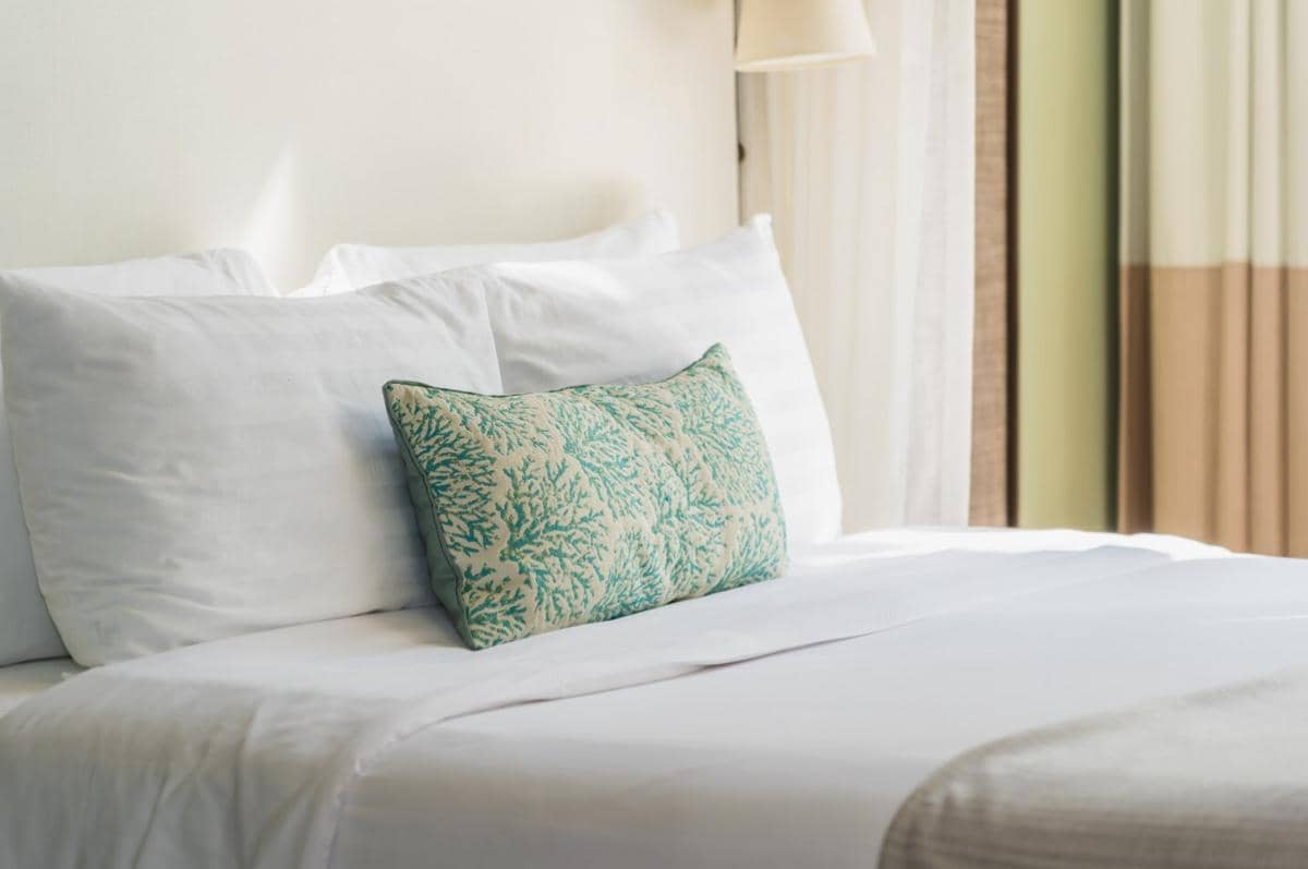 Tempat tidur dengan sprei linen putih lembut dan bantal bermotif hijau di kamar bernuansa hangat dengan tirai terbuka.