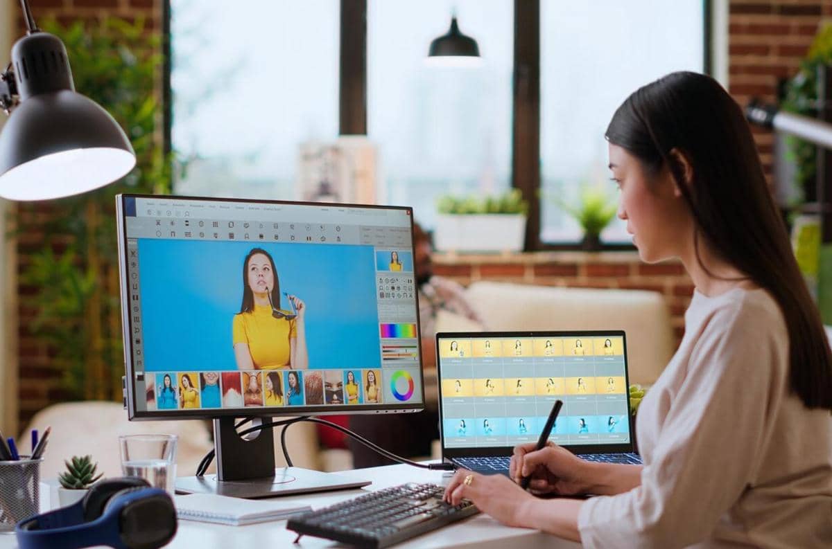 Seorang desainer grafis bekerja di depan komputer dengan tablet pen stylus, mengedit foto di ruang kerja modern yang terang.