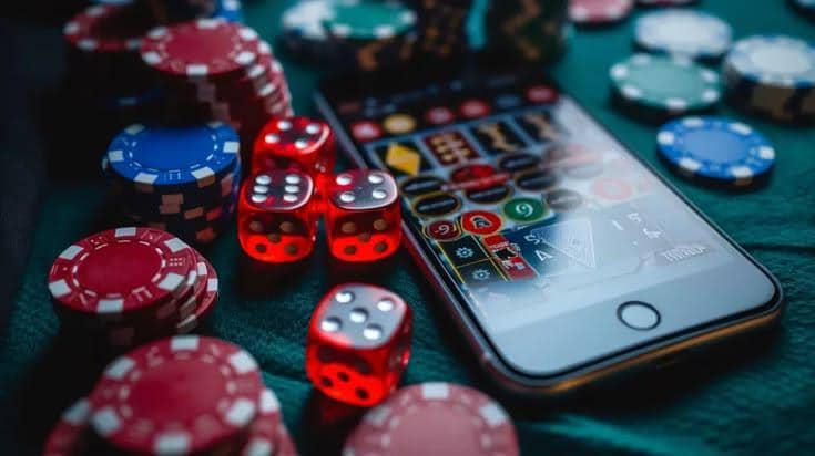 Smartphone menampilkan aplikasi judi online dikelilingi chip kasino dan dadu di atas meja permainan.