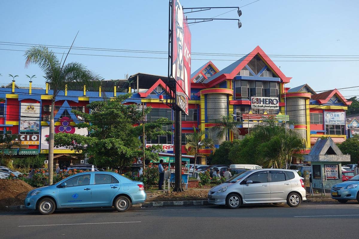 Bagian luar pusat perbelanjaan berwarna cerah di Mataram dengan beberapa mobil terparkir di tepi jalan pada siang hari.