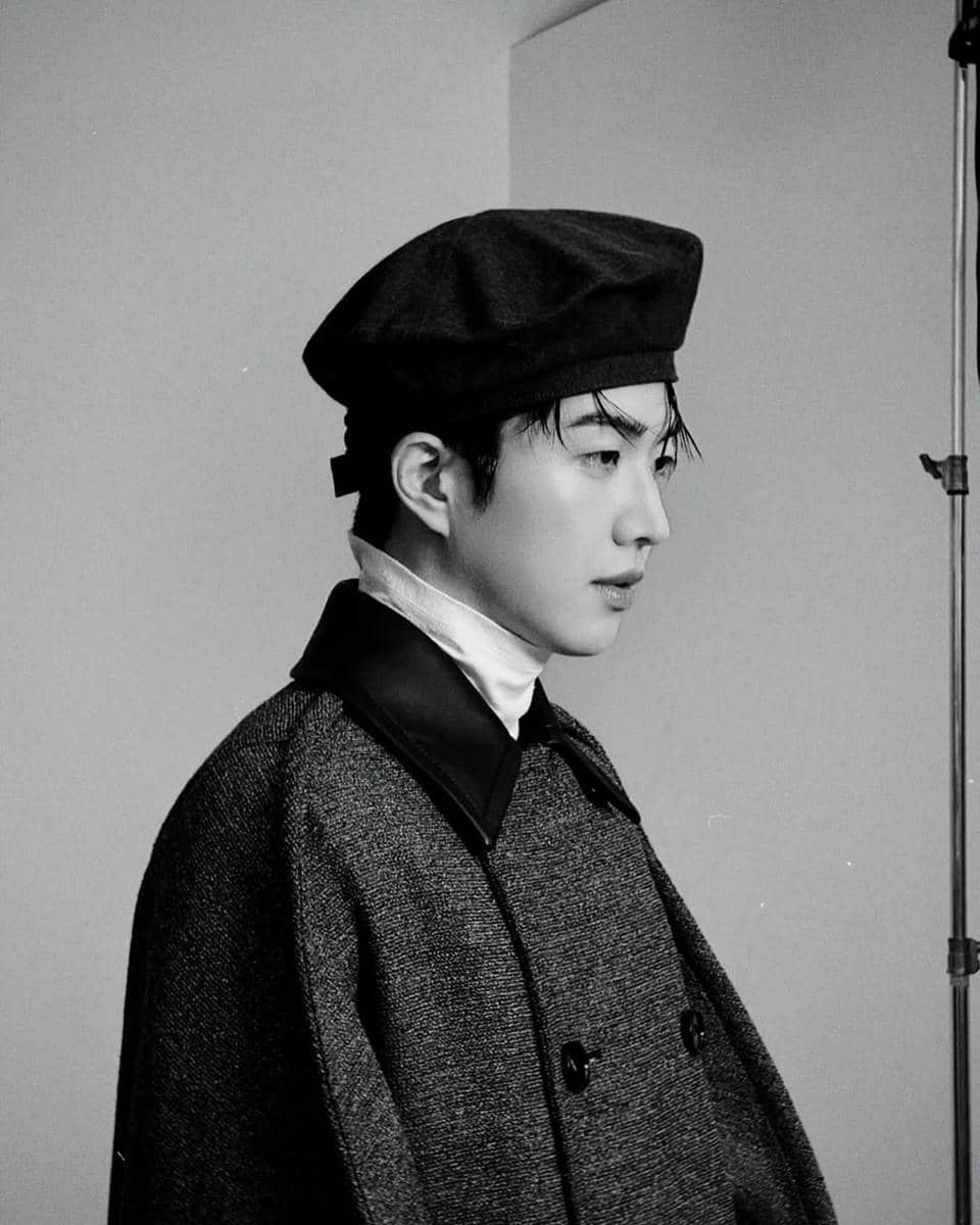 Seorang pria mengenakan mantel tebal dan topi baret hitam berpose dalam foto hitam putih dengan latar sederhana di studio.