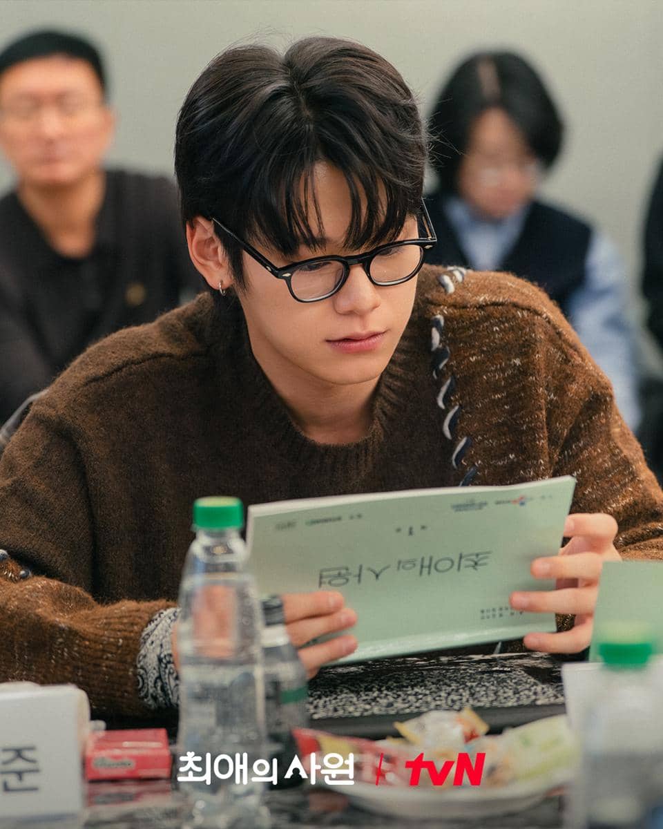 Seorang aktor mengenakan kacamata dan sweater cokelat sedang membaca naskah drama My Bias, My Boss di sesi pembacaan naskah tvN.