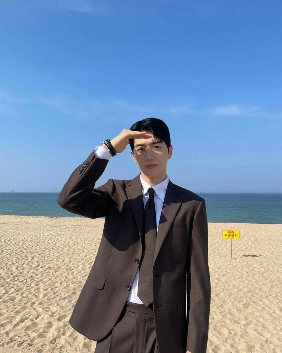Seorang pria mengenakan setelan jas cokelat dan dasi hitam berdiri di pantai berpasir dengan latar belakang laut biru dan langit cerah.