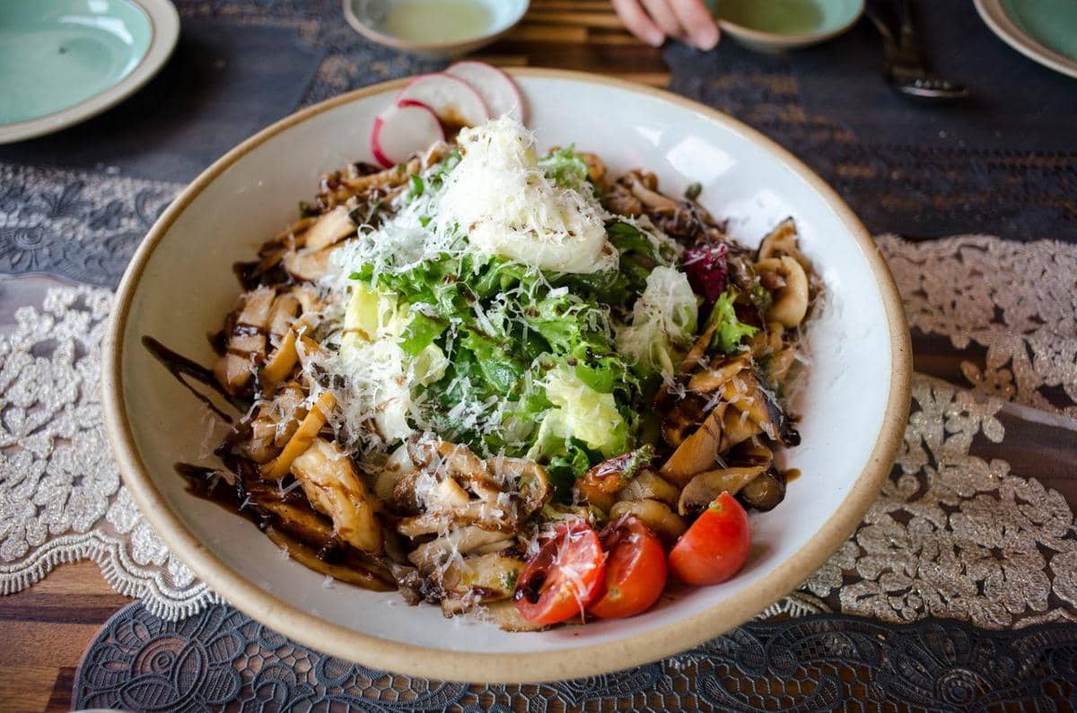 Semangkuk salad jamur dengan parutan keju, sayuran hijau segar, tomat ceri, dan saus balsamik di atas meja makan.
