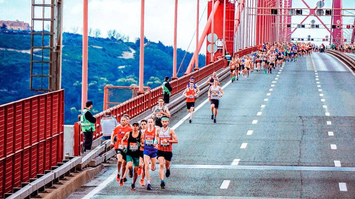 Para pelari mengikuti lomba maraton di atas jembatan besar berwarna merah dengan latar perbukitan hijau di kejauhan.