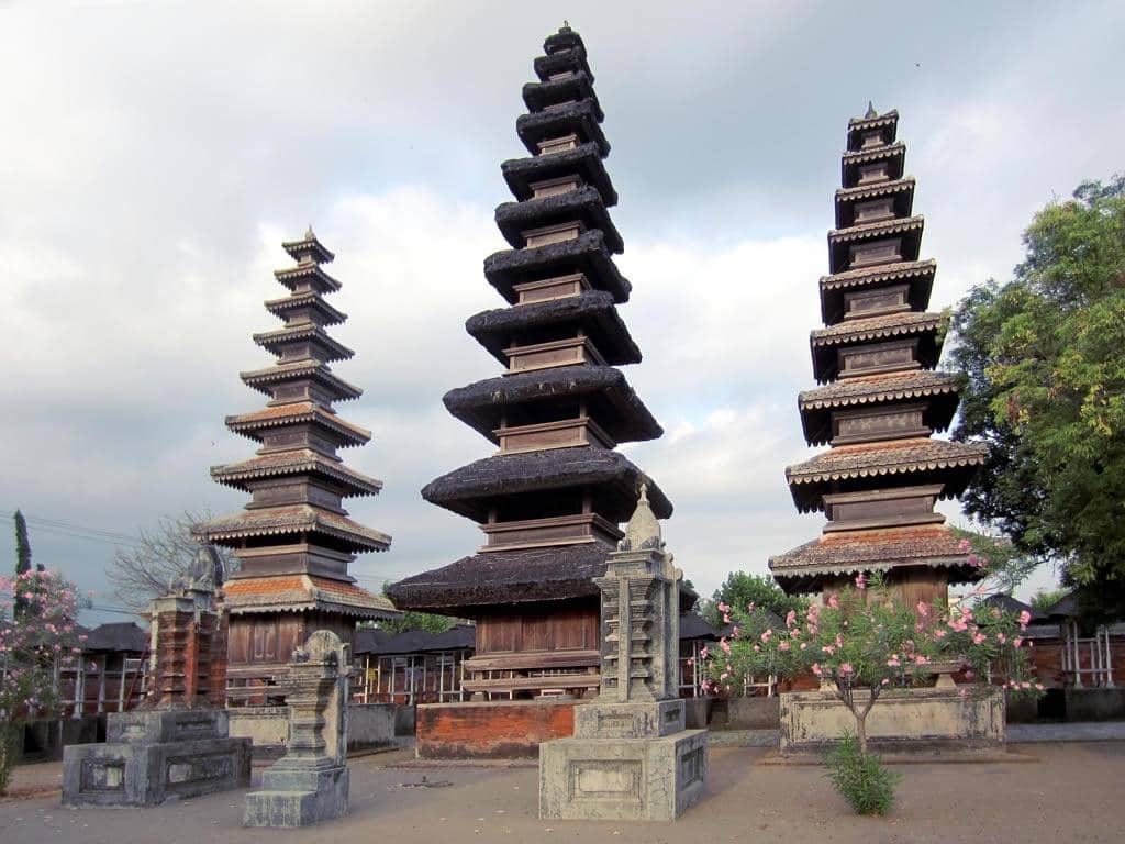 Tiga menara bertingkat khas arsitektur Bali di Pura Meru, Mataram, dengan langit berawan dan pepohonan di sekitarnya.
