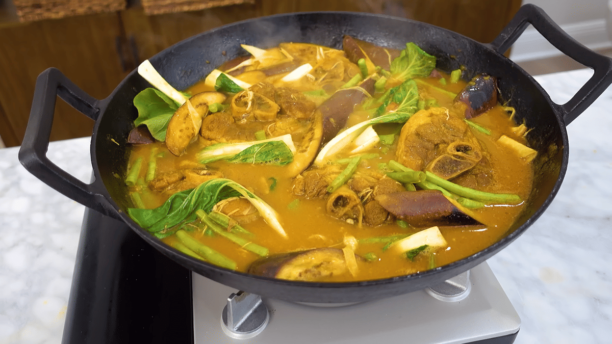 Kare-kare dalam wajan besar berisi potongan daging sapi, terong, kacang panjang, dan sayuran hijau dengan kuah kacang berwarna oranye.