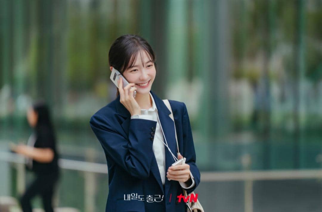 Seorang wanita mengenakan setelan jas biru tersenyum sambil berbicara di telepon di area luar gedung kaca modern.
