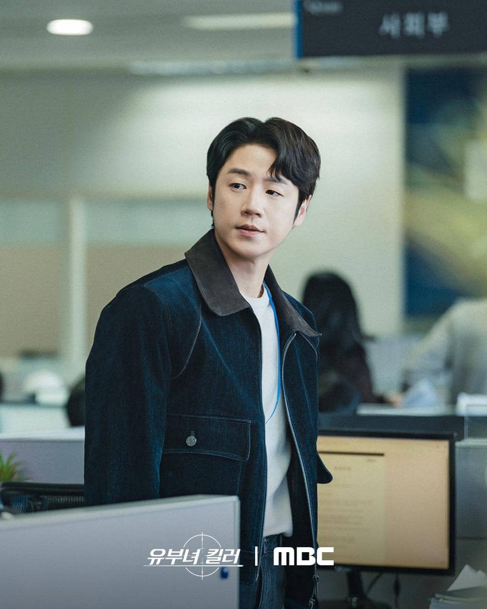 Seorang pria mengenakan jaket denim gelap berdiri di area kantor dengan komputer dan meja kerja di sekitarnya.