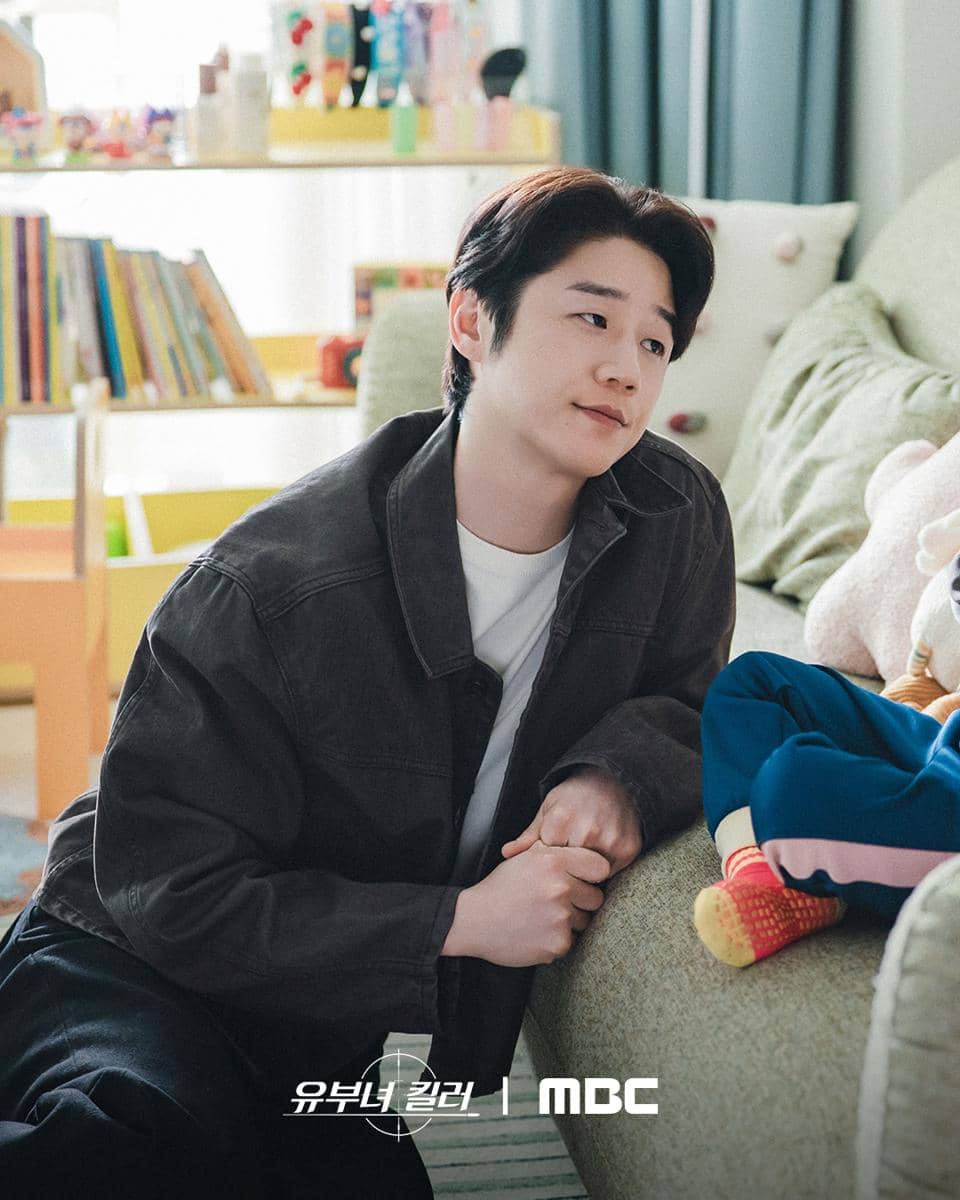Seorang pria mengenakan jaket gelap duduk di lantai sambil menatap seseorang di sofa dalam adegan drama Killer of Married Woman.