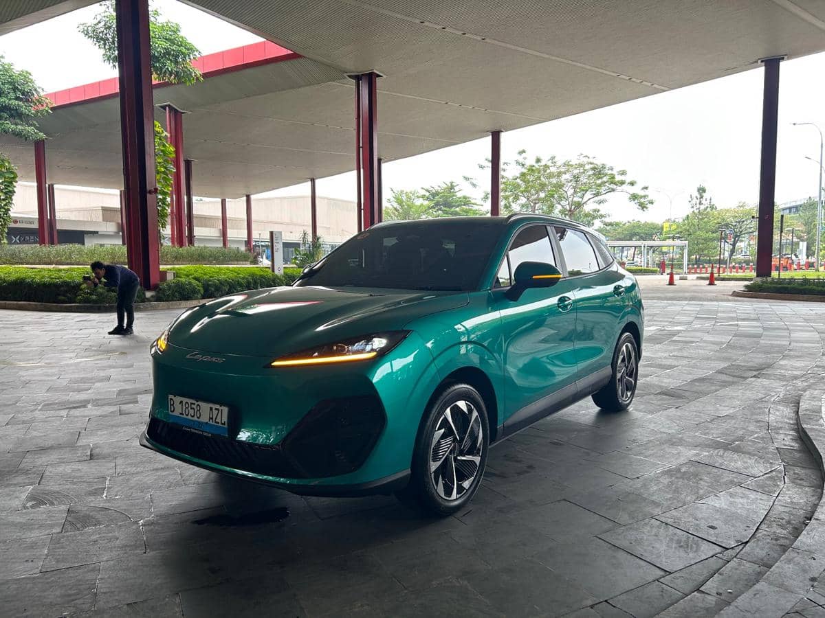 Mobil SUV berwarna hijau toska dengan desain modern diparkir di area luar gedung beratap lebar dengan tiang merah dan pepohonan di sekitarnya.