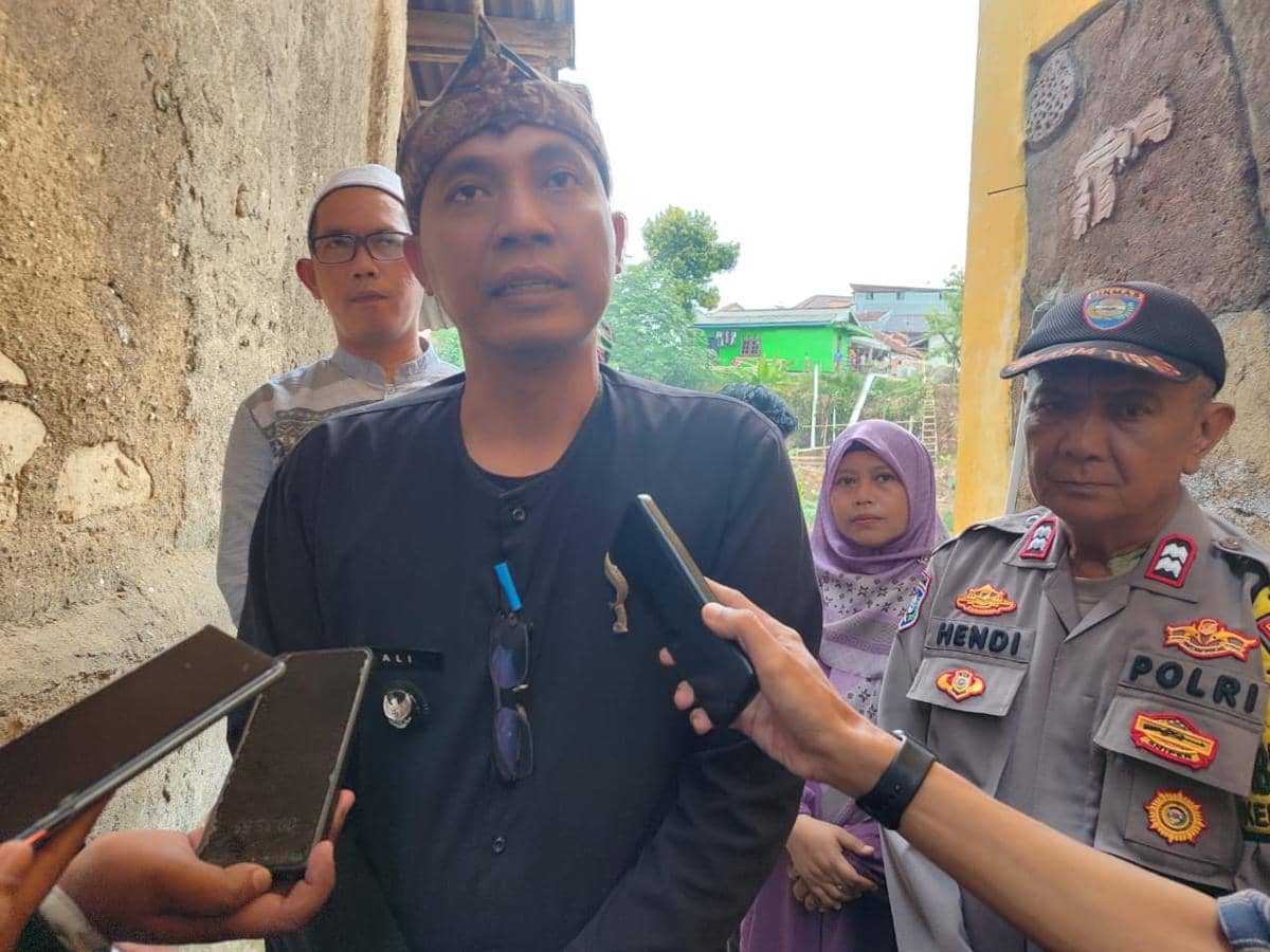 Seorang lurah mengenakan pakaian hitam diwawancarai oleh beberapa jurnalis di sebuah gang sempit bersama petugas polisi dan warga.