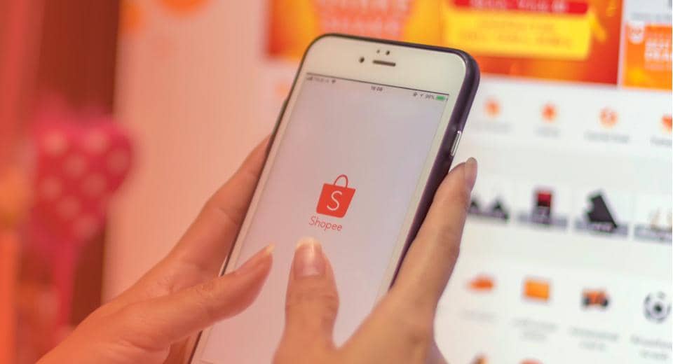 Seseorang memegang ponsel yang menampilkan aplikasi Shopee dengan latar belakang layar komputer berisi halaman Shopee.
