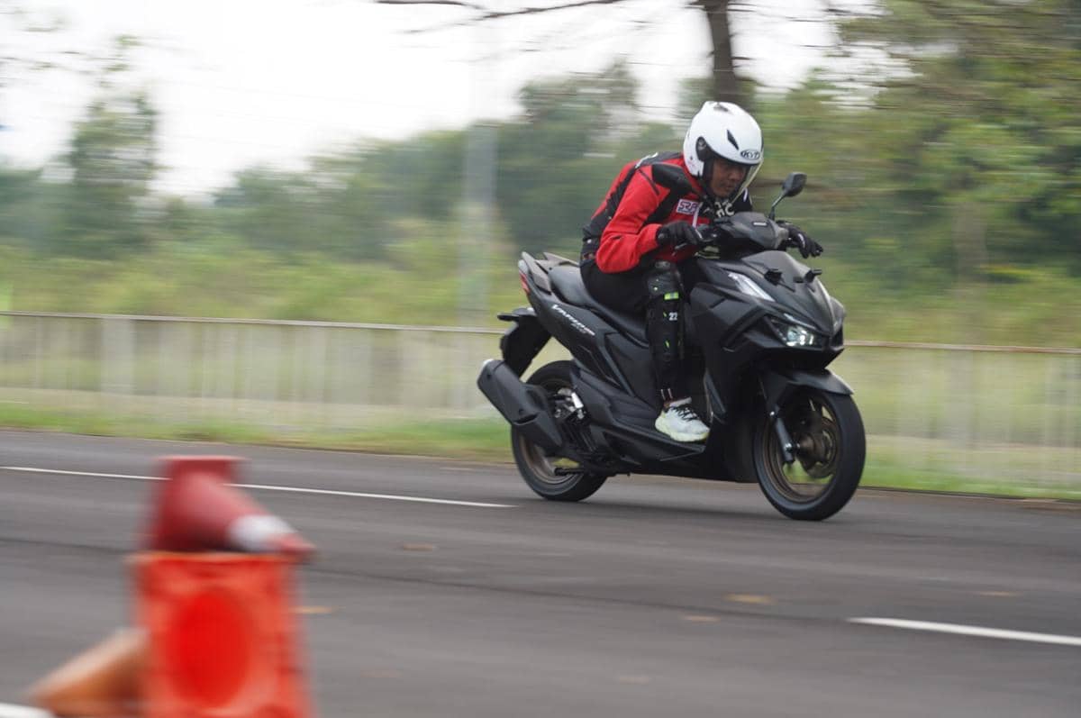 Seorang pengendara dengan jaket merah dan helm putih mengendarai motor skuter hitam di jalan aspal dengan kecepatan tinggi.