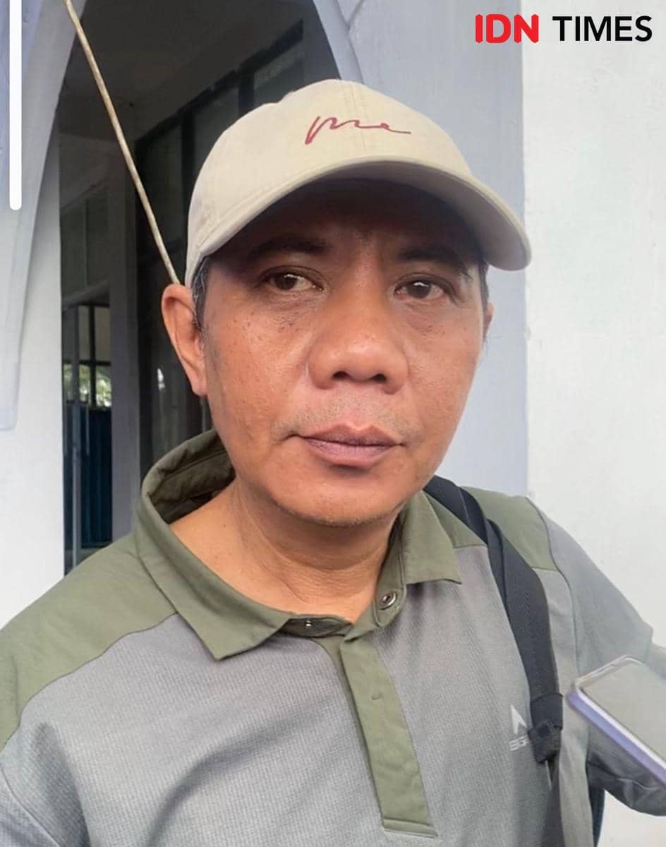 Seorang pria mengenakan topi krem dan kemeja abu-abu hijau sedang berbicara di luar ruangan dengan latar dinding putih.