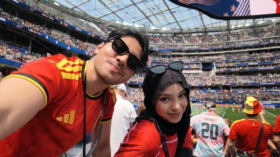 Dua penonton mengenakan jersey merah berfoto selfie di stadion besar yang dipenuhi penonton saat pertandingan Piala Dunia 2026.