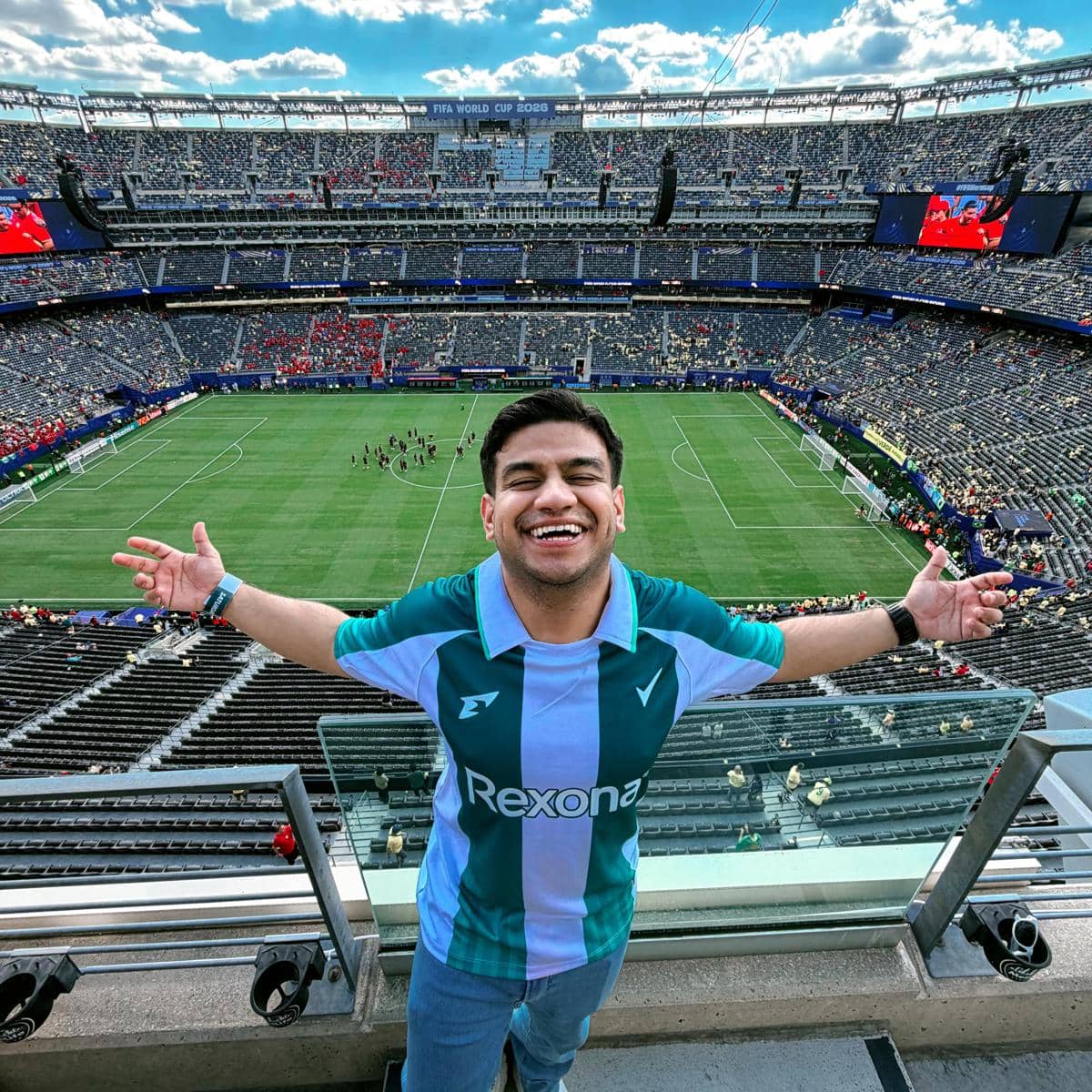 Seorang pria mengenakan jersey hijau putih tersenyum lebar dengan tangan terbuka di stadion besar saat pertandingan Piala Dunia 2026.