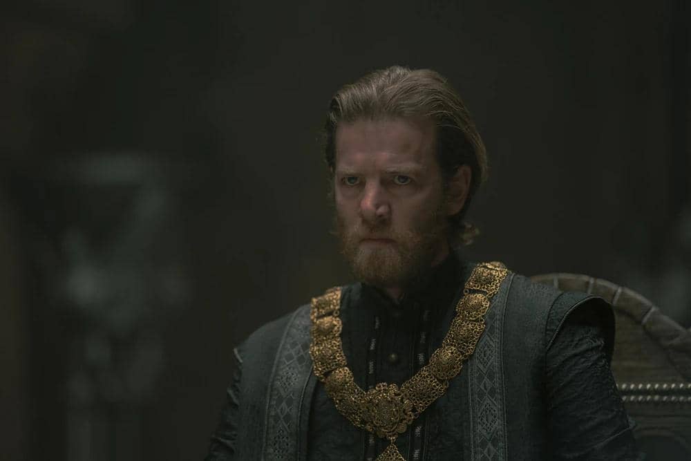 Seorang pria berambut pirang berjanggut mengenakan pakaian gelap berhias rantai emas duduk di kursi dalam adegan House of the Dragon Season 3.