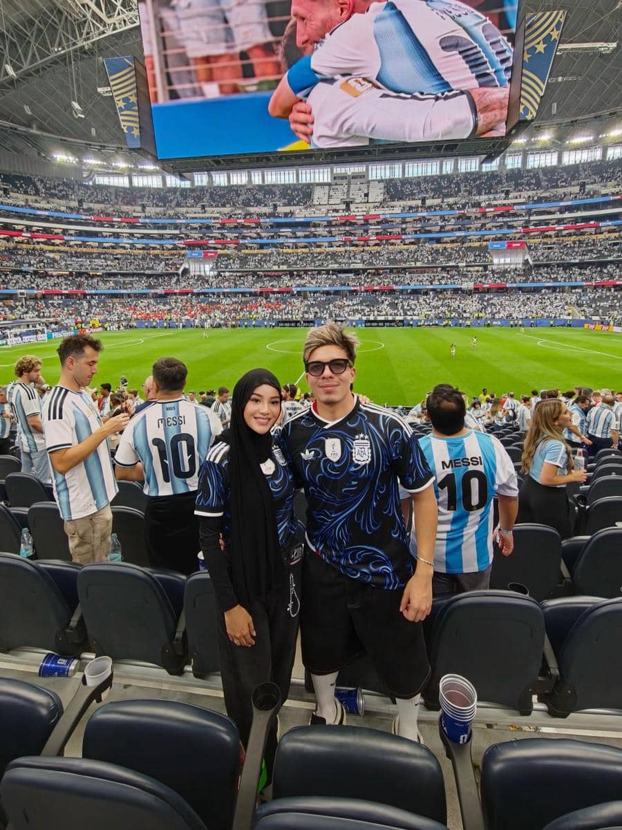 Sepasang penonton mengenakan jersey tim Argentina berpose di tribun stadion besar saat pertandingan fase grup Piala Dunia 2026.