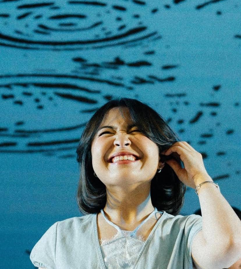 Seorang perempuan berambut pendek tersenyum lebar sambil memegang rambutnya dengan latar belakang pola air berwarna biru.