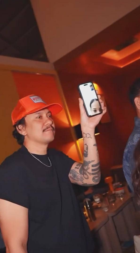 Seorang pria mengenakan topi oranye dan kaus hitam sedang melakukan panggilan video menggunakan ponsel di pesta perpisahan Indonesian Idol Season 14.