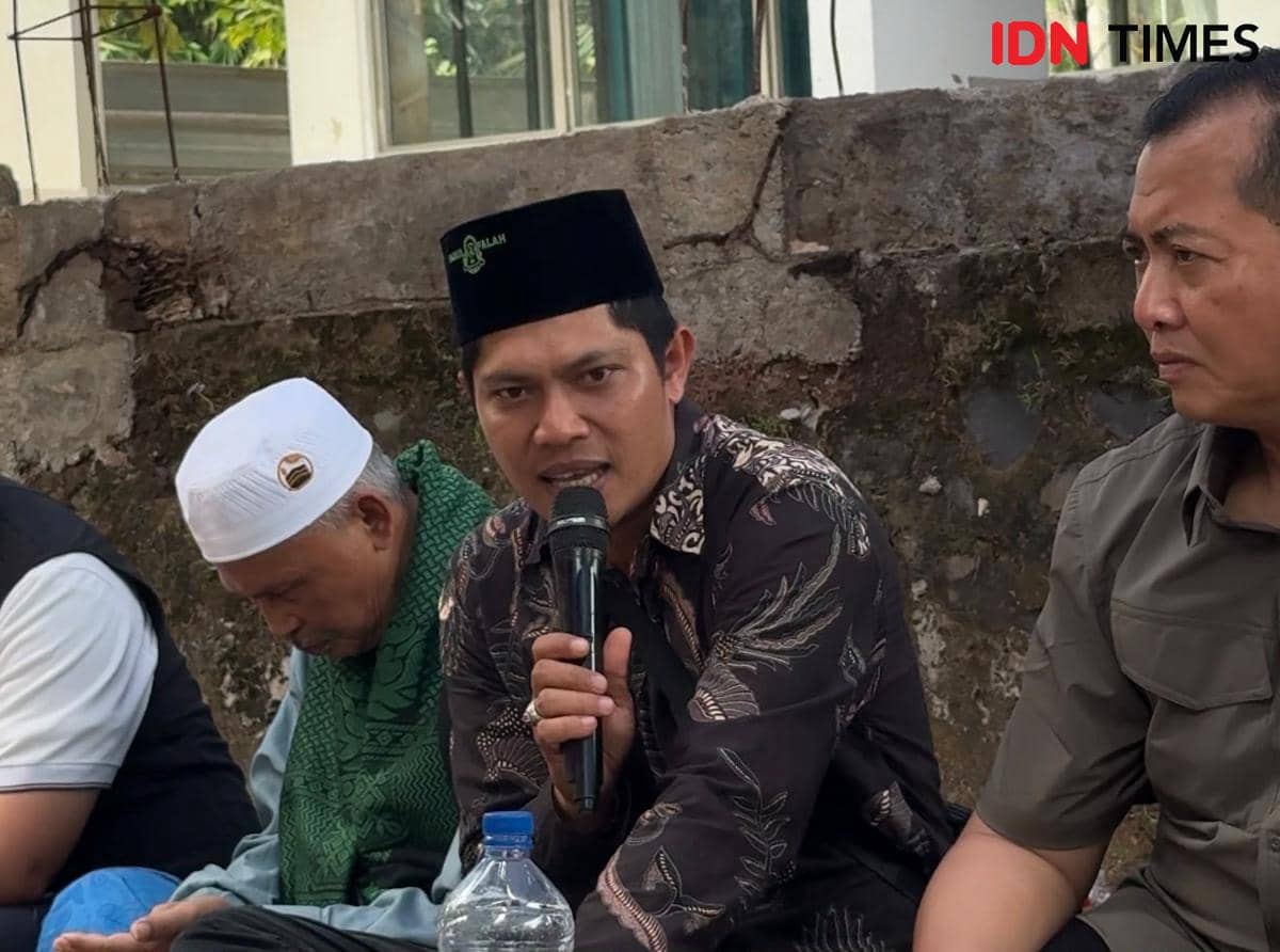 Seorang pria mengenakan batik hitam dan peci hitam berbicara menggunakan mikrofon di samping dua pria lain dalam acara luar ruangan.